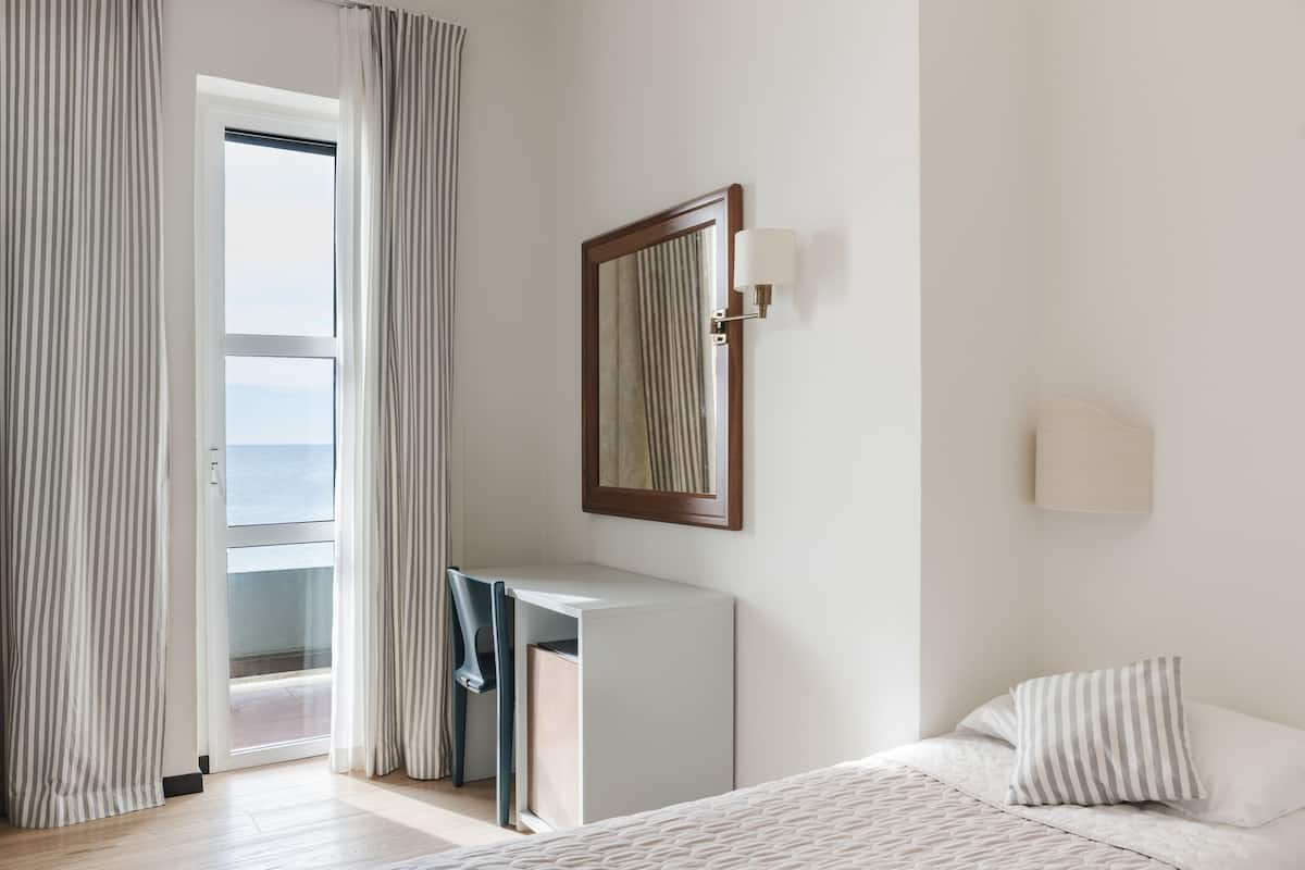 A bedroom with a bed, a desk, and a chair. There is a window with curtains, a wall-mounted light, and a view of the outdoors.