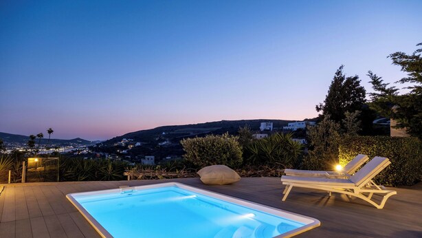 A rectangular swimming pool with a light blue tint, surrounded by a wooden deck. There are two lounge chairs with cushions and a large rock in the center. The background features a mountainous landscape with buildings and a clear sky. - Azzurro Bianco Suites (Paros)