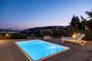 A rectangular swimming pool with a light blue tint, surrounded by a wooden deck. There are two lounge chairs with cushions and a large rock in the center. The background features a mountainous landscape with buildings and a clear sky.