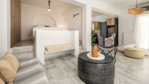 A modern living room with a sofa, a round black ottoman, a chair, and a bed with a white headboard. - Azzurro Bianco Suites (Paros)