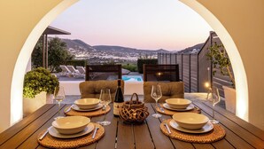 An outdoor dining area with a view of a mountainous landscape, a pool, and lounge chairs. - Azzurro Bianco Suites (Paros)