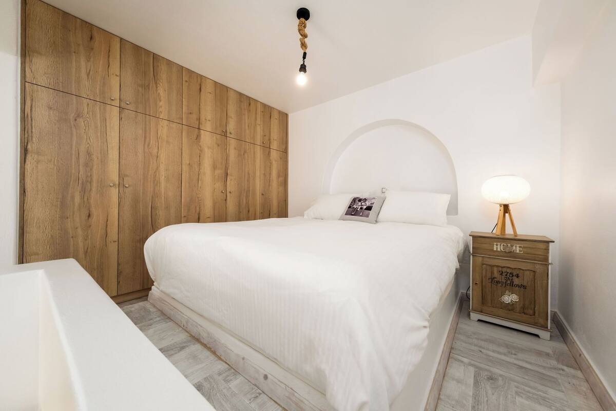 Un dormitorio con un armario de madera, una cama con ropa blanca, una mesita de noche con lámpara y un pequeño aparador.
