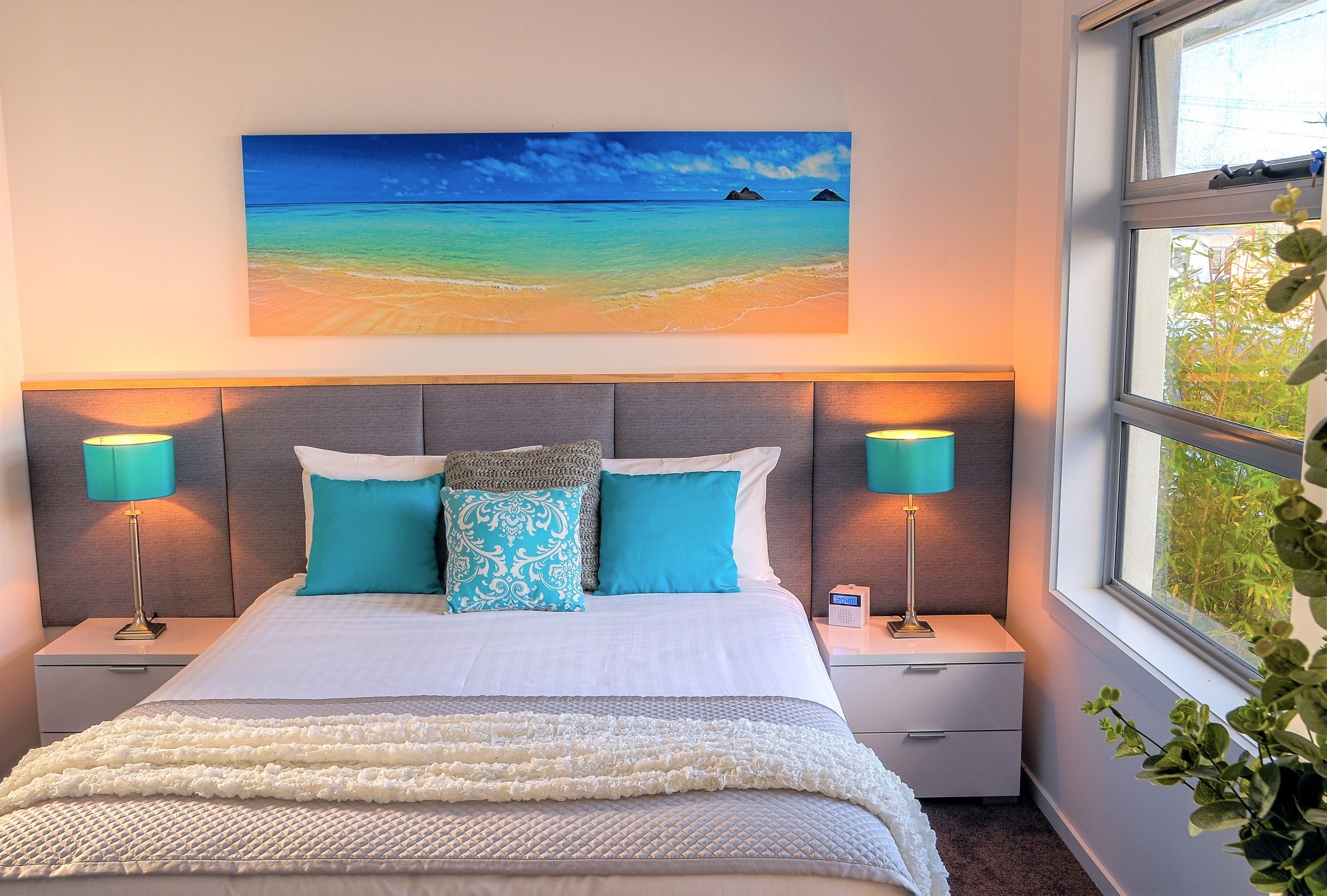 A bedroom with a bed, two bedside tables, a nightstand, a painting of a beach, and a window with a view of greenery.