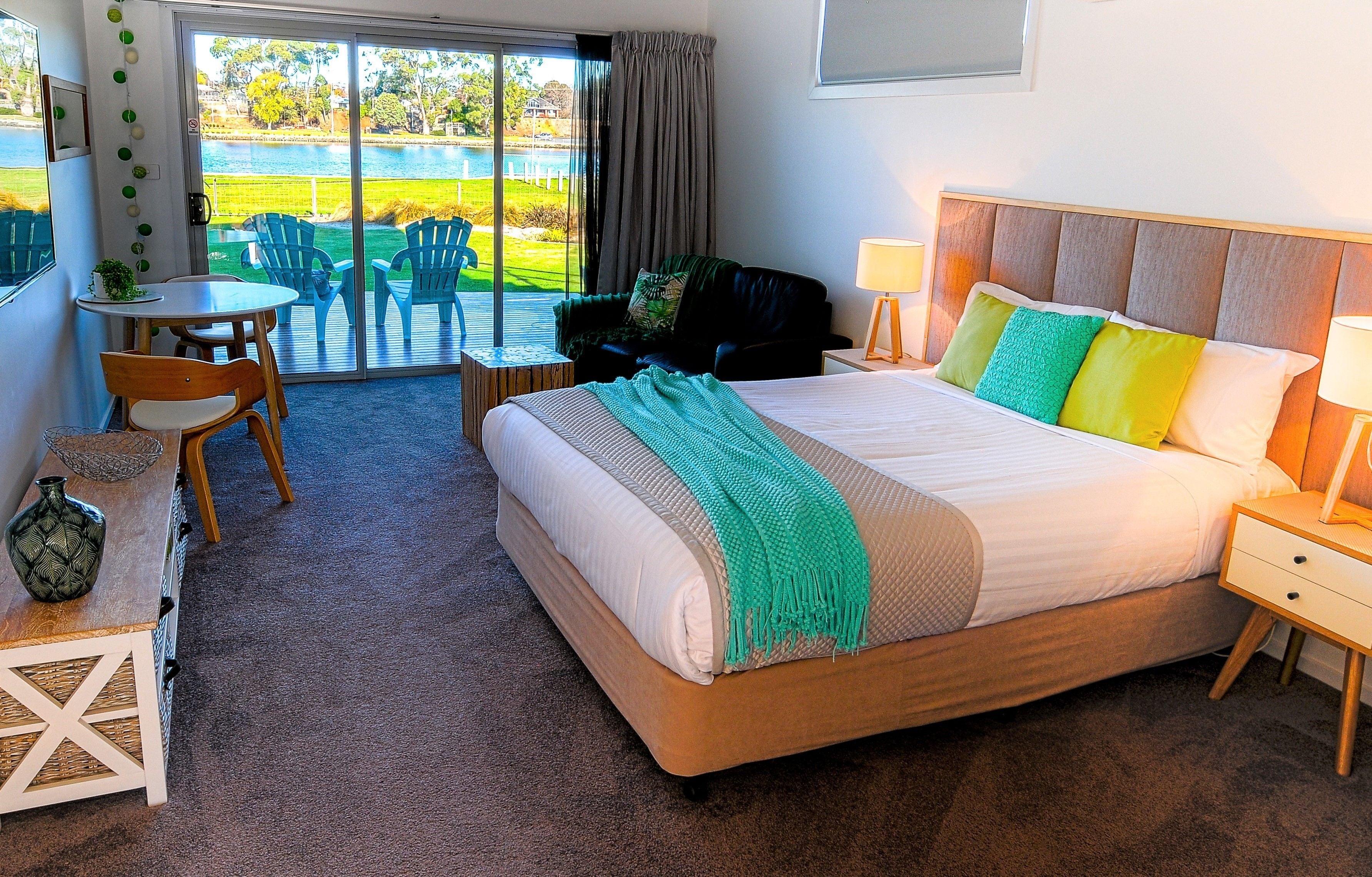 A hotel room with a large bed, a desk, a chair, a small table, and a view of a lake through the sliding glass door.