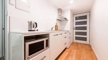 Cocina compacta con armarios blancos, microondas, hervidor y tabla de cortar de madera.