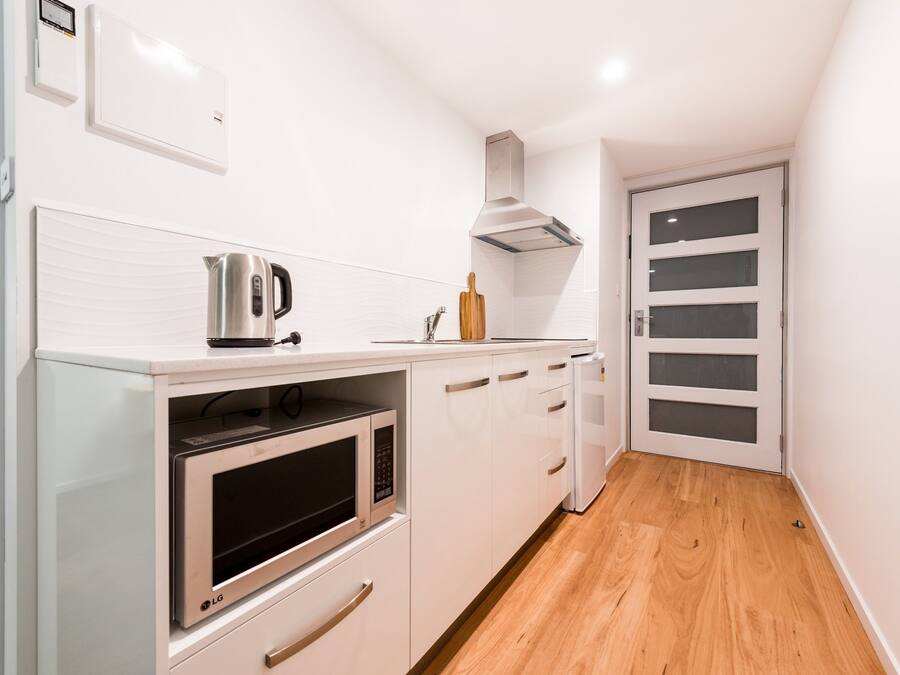 Cocina compacta con armarios blancos, microondas, hervidor y tabla de cortar de madera.