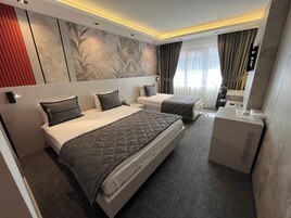 Quarto de hotel com duas camas, uma escrivaninha, um espelho e uma janela grande com cortinas.