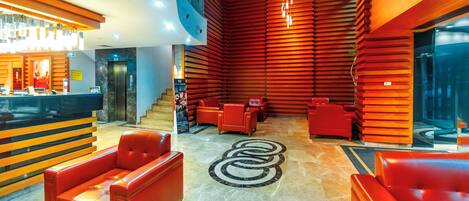 Un vestíbulo de hotel con mostrador de recepción, asientos rojos, una escalera y una moderna lámpara de araña.