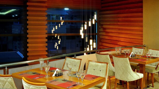 Un espace repas avec des tables et des chaises en bois, un lustre moderne et une vue dégagée sur l’extérieur grâce à une grande fenêtre.