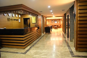 Uma área de recepção com um balcão de madeira, vários relógios e um bar visível ao fundo.