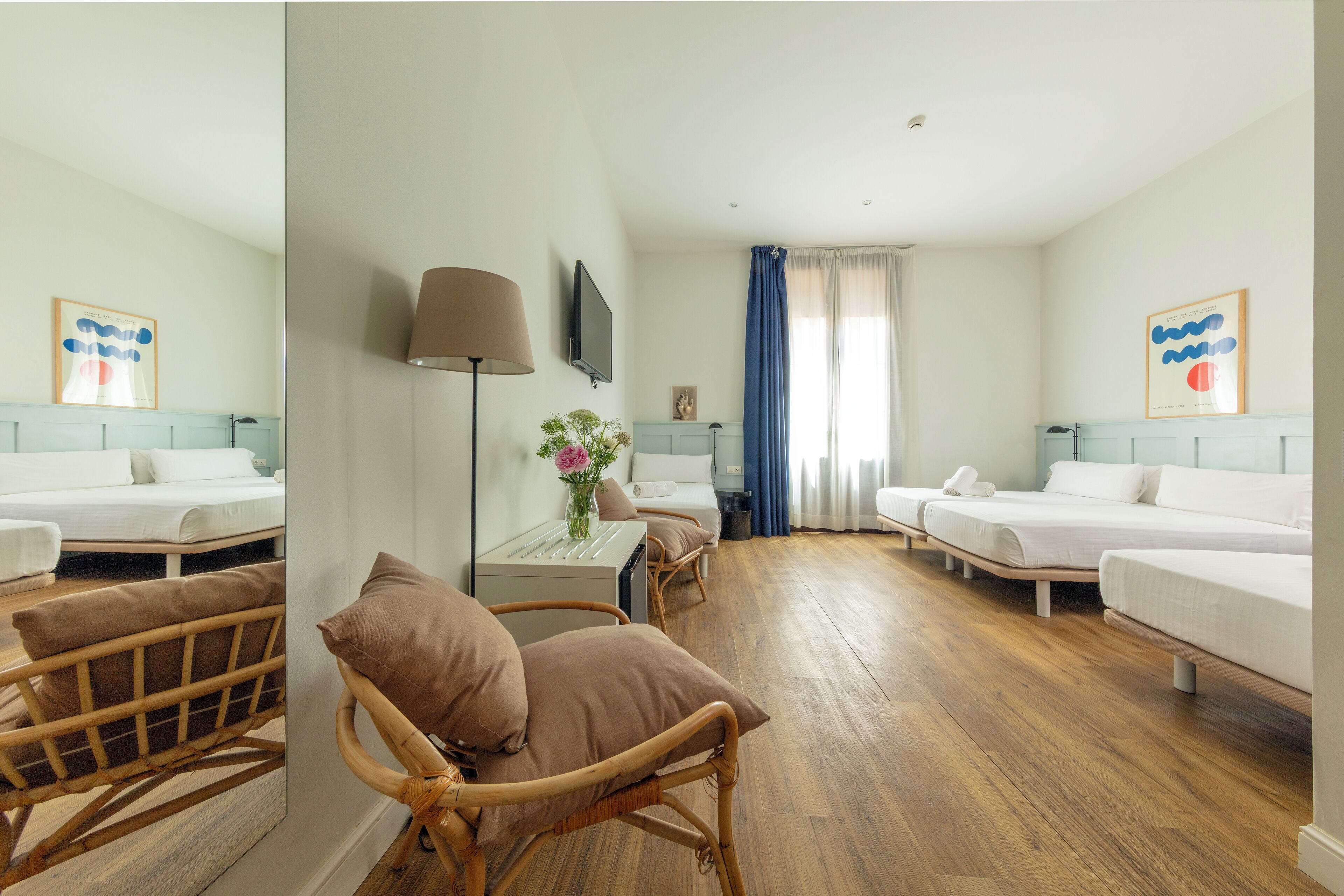 Habitación de hotel con dos camas individuales, una zona de estar con una silla y una mesa, un televisor montado en la pared y un ventanal con cortinas.