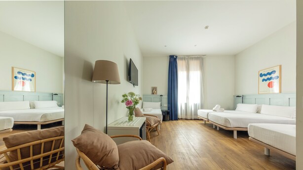 Une chambre d’hôtel avec deux lits simples, un coin salon comprenant une chaise et une table, un téléviseur fixé au mur et une grande fenêtre avec des rideaux.