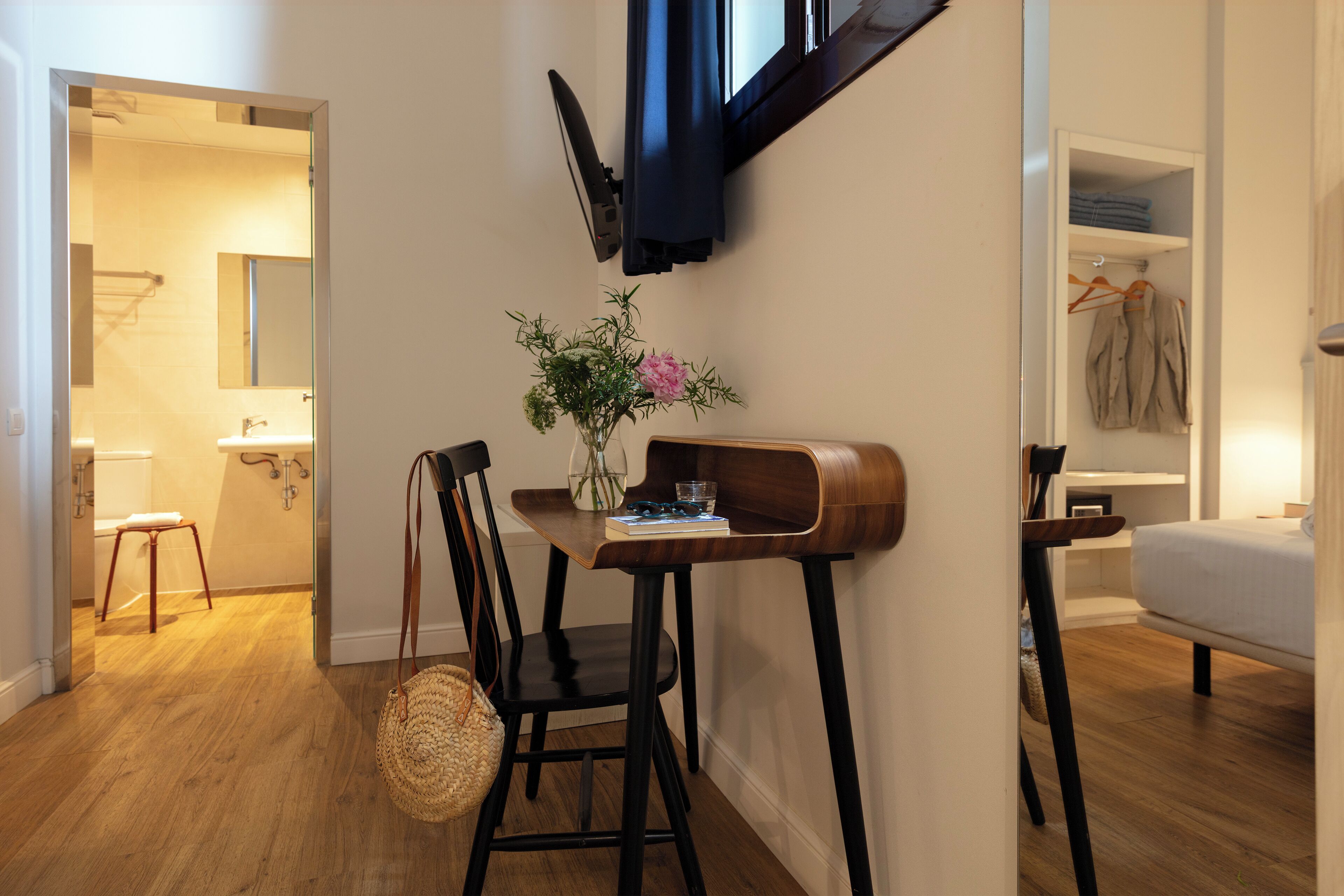 Un baño moderno con inodoro, lavamanos y perchero para toallas. Se ve una silla de madera con un bolso tejido y un florero con flores.