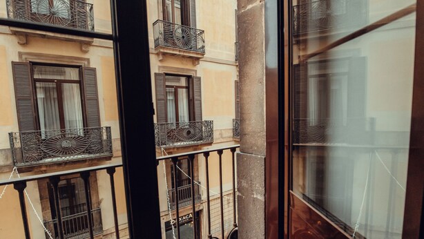 Vista a través de un ventanal de un edificio con varios balcones, cada uno con barandillas de metal y ventanas con contraventanas.