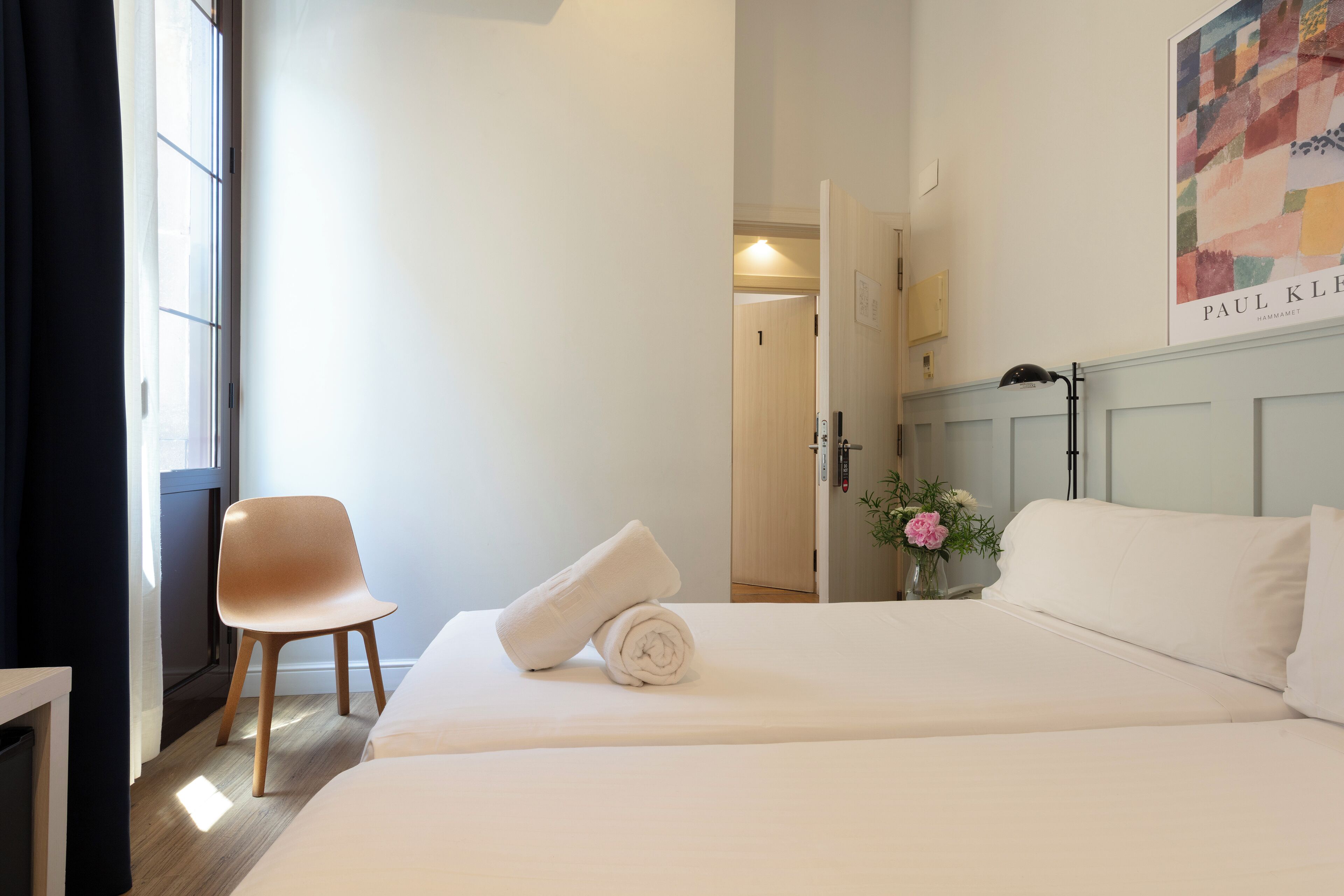 Una habitación de hotel con cama, una silla, ventana con cortinas y un cartel en la pared.