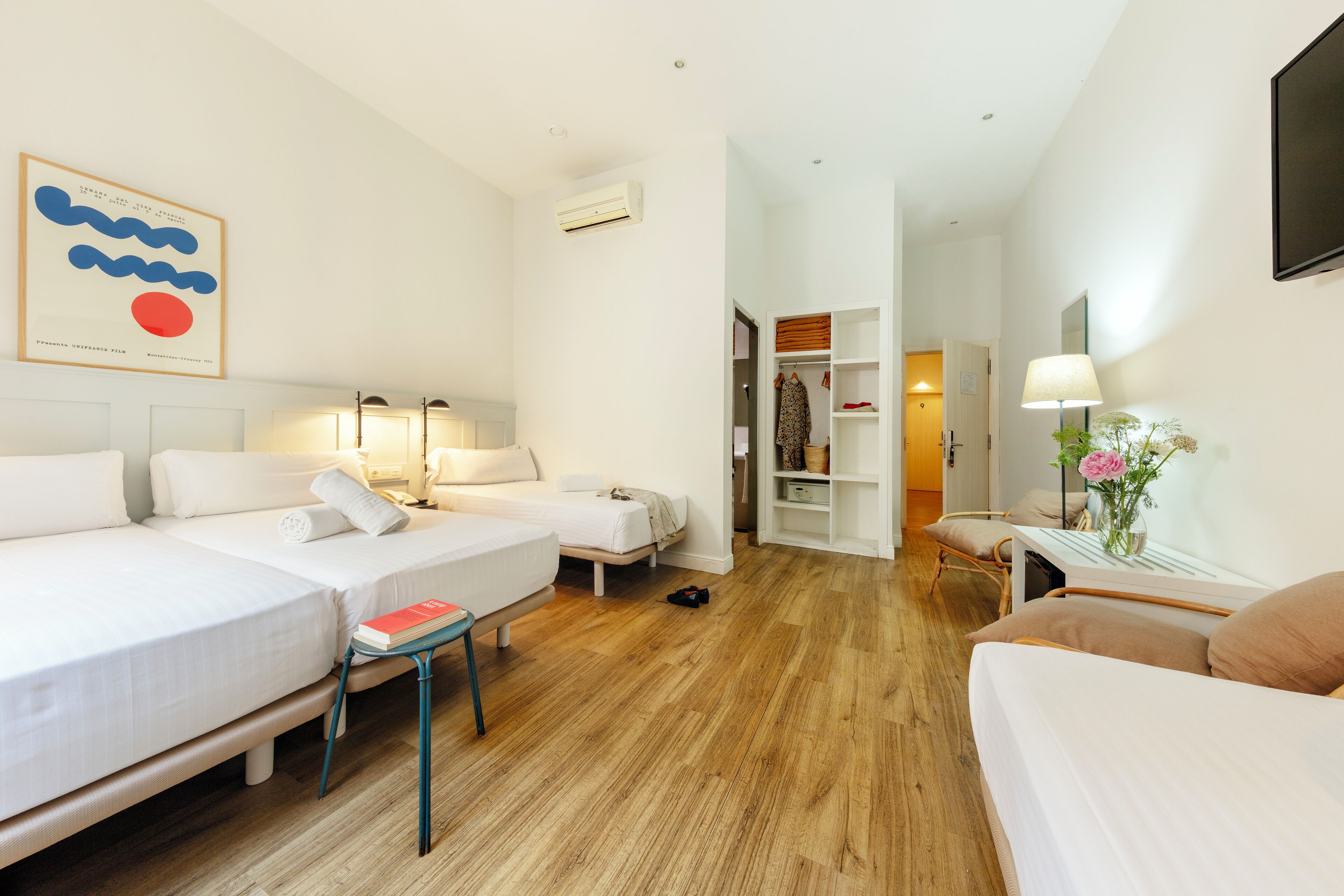 Habitación de hotel con dos camas, un escritorio con lámpara, un armario y un televisor en la pared.