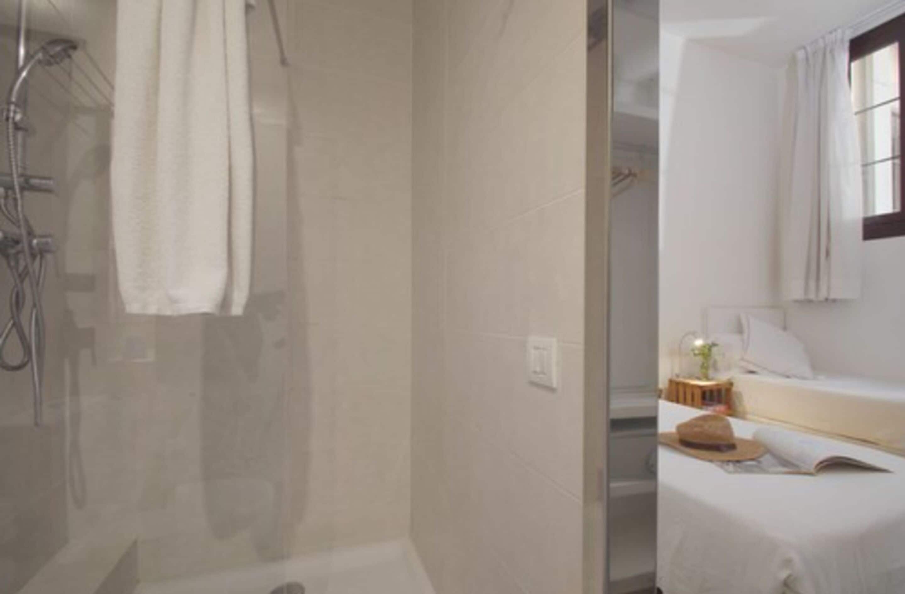 Un baño con cabina de ducha de vidrio, una toalla colgada en la pared y una cama con mesita de noche en la habitación contigua.