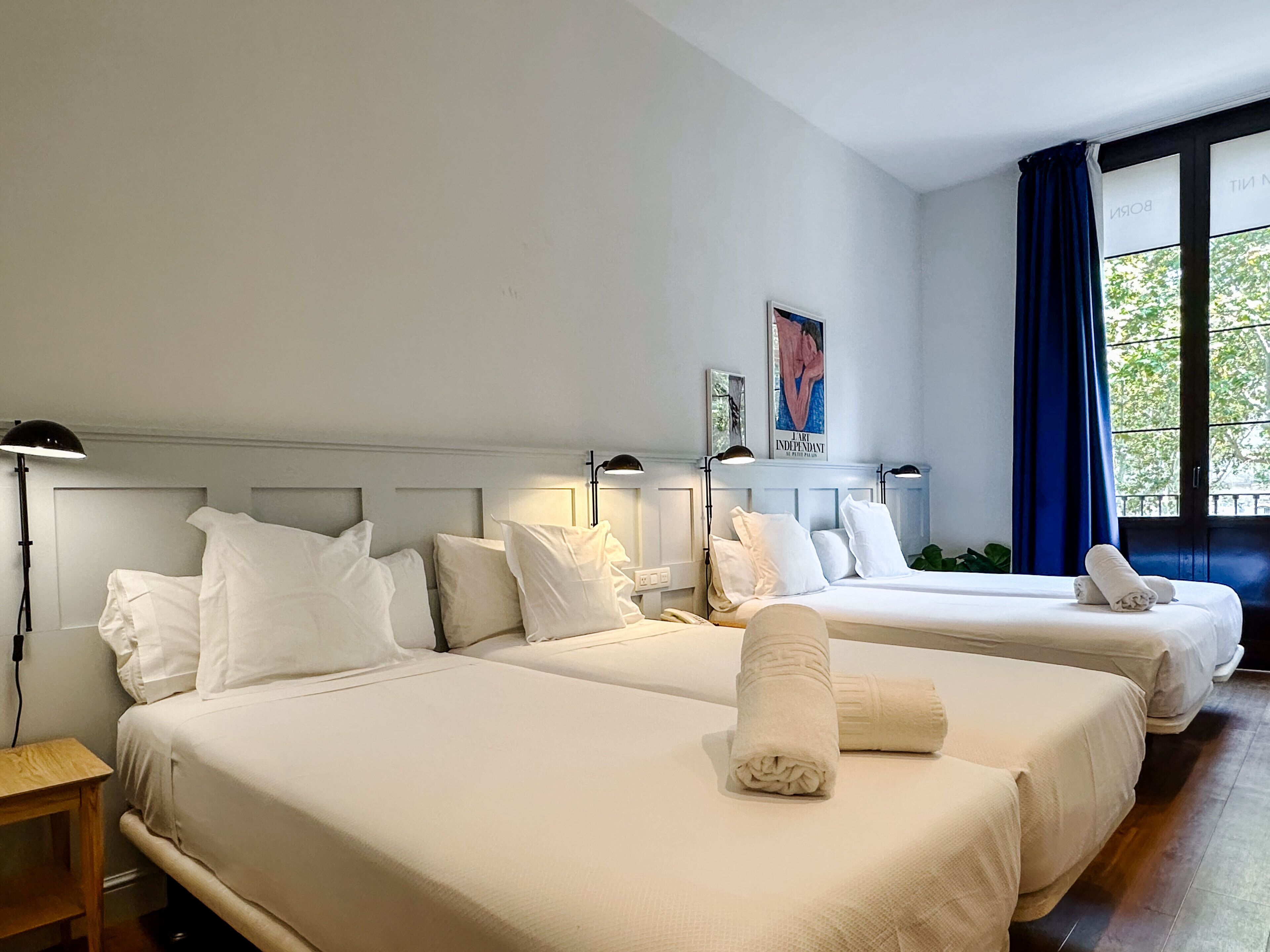 Hotelkamer met twee bedden, een nachtkastje, een lamp, een poster aan de muur en een raam met gordijnen.