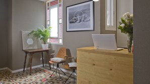 A reception area with a wooden desk, a laptop, a framed photograph, a potted plant, and a chair. - Som Nit Triomf (Barcelona)