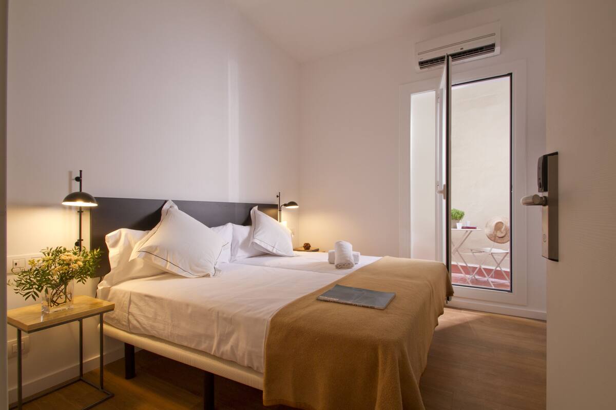 Una habitación de hotel con cama, mesita de noche, lámpara y vistas al balcón exterior.