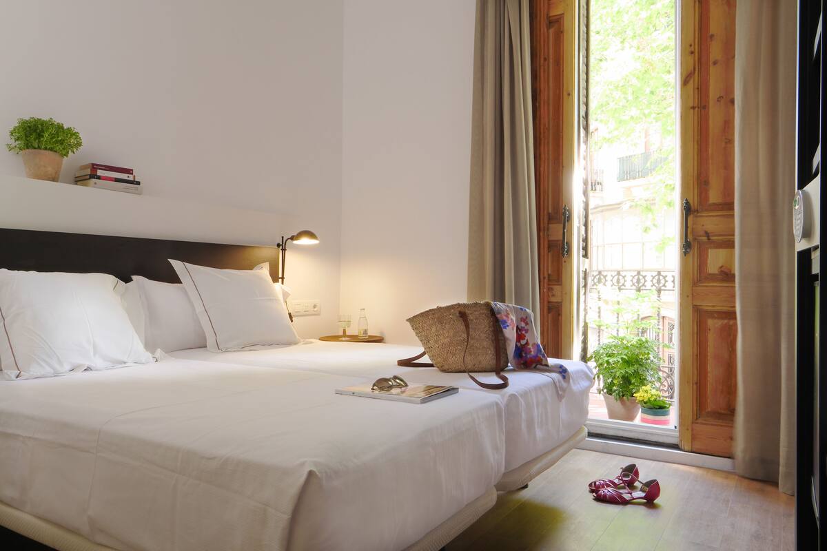 Una cama bien hecha con sábanas blancas, una lámpara de noche, una mesita con un vaso y una planta en maceta en el alféizar.