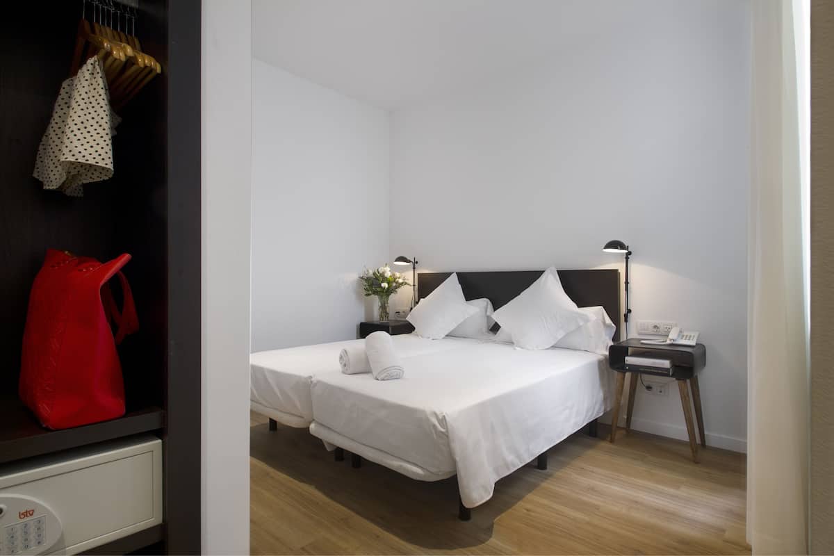 Una cama bien hecha con ropa de cama blanca, una mesita de noche con lámpara y un pequeño mueble con un teléfono.