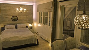 A bedroom with a bed, bedside lamps, a chandelier, a dining table with chairs, and a window with blinds. - Apartments Huili (Kuopio)