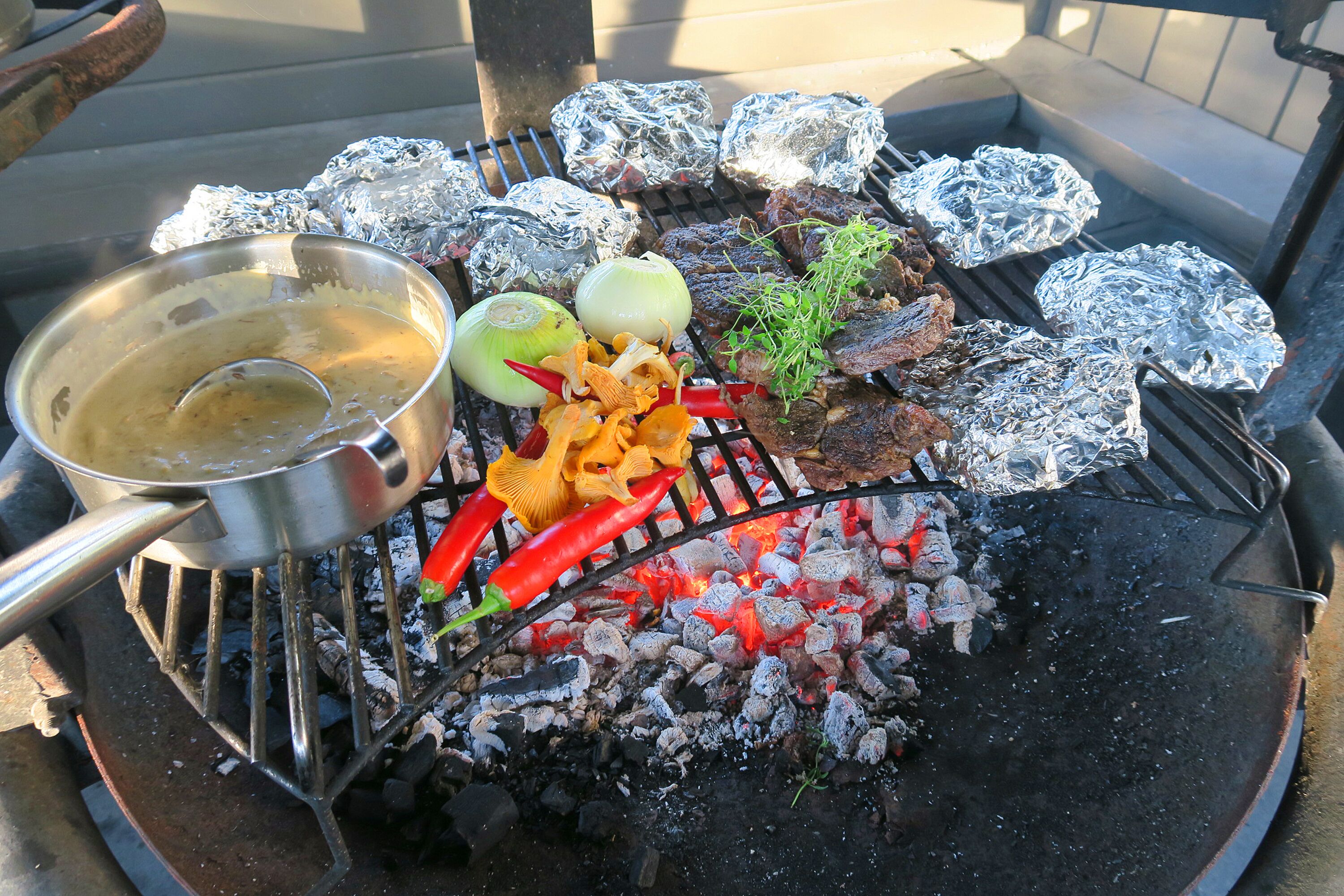 Una parrilla con alimentos envueltos en papel de aluminio, una olla y verduras.