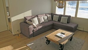 A modern living room with a sectional sofa, a wooden coffee table with wheels, and a view of a snowy landscape through large windows. - Apartments Huili (Kuopio)