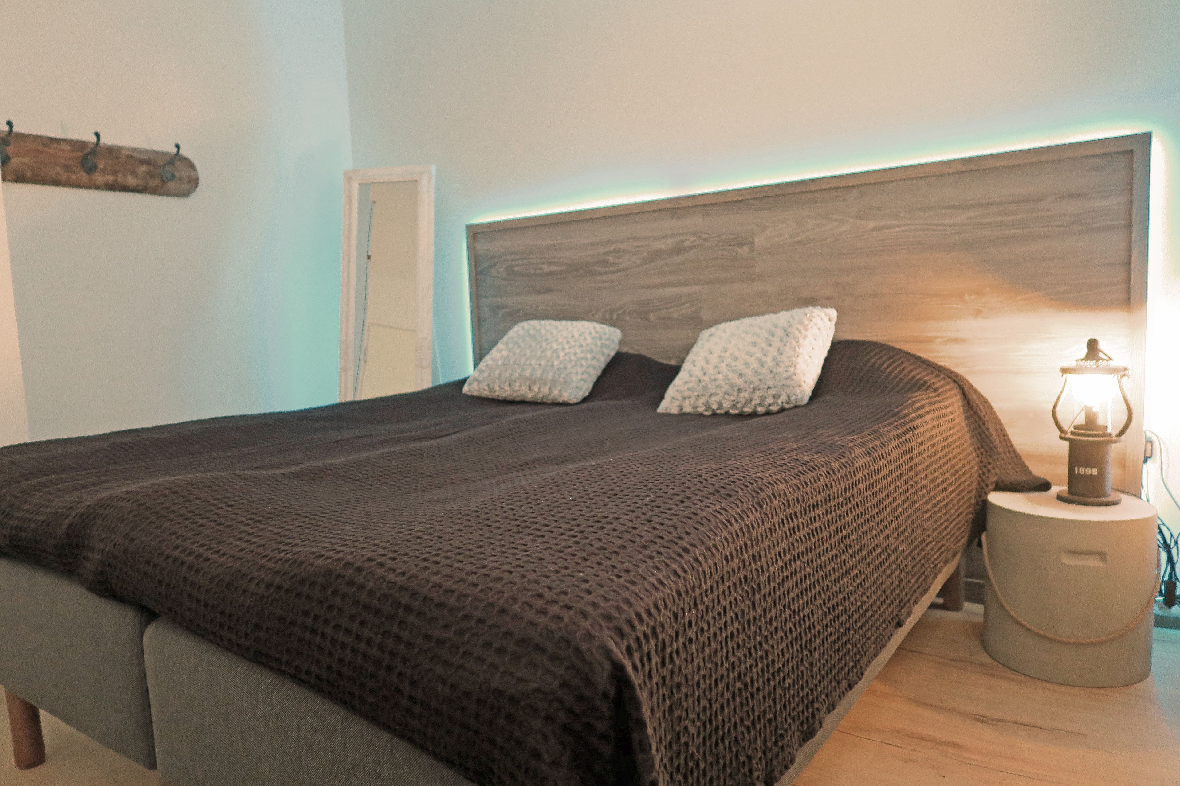 A bedroom with a wooden headboard, a bed with a brown blanket, two pillows, a bedside table with a lamp, and a coat rack.