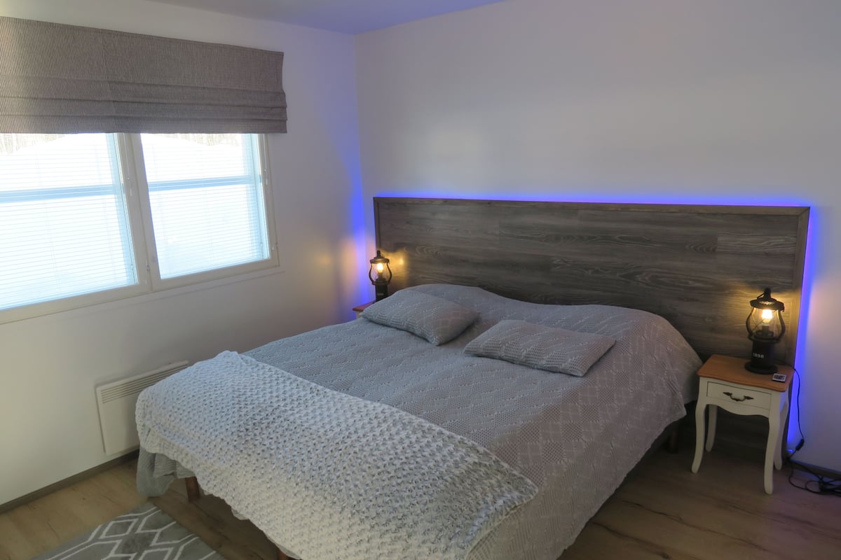 A bedroom with a bed, two bedside lamps, a nightstand, a window with blinds, and a wooden headboard.