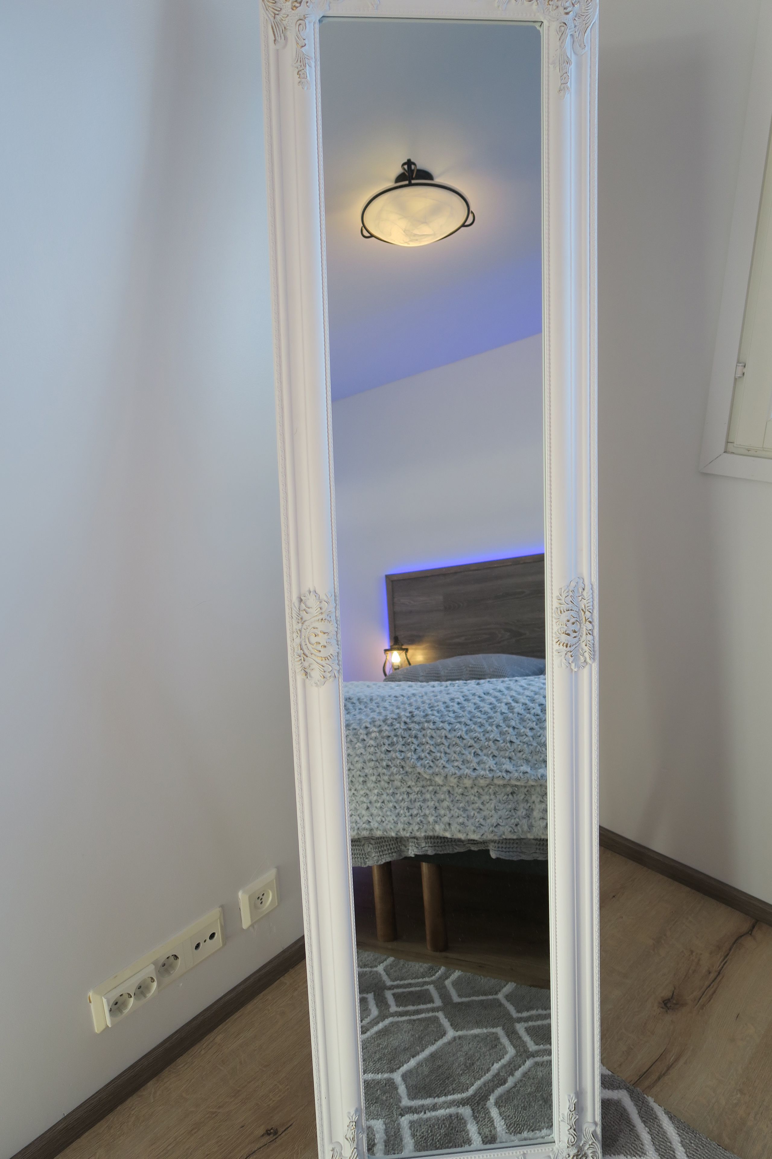 A mirror with ornate detailing, a bed with a patterned bedspread, a wall socket, and a patterned rug.