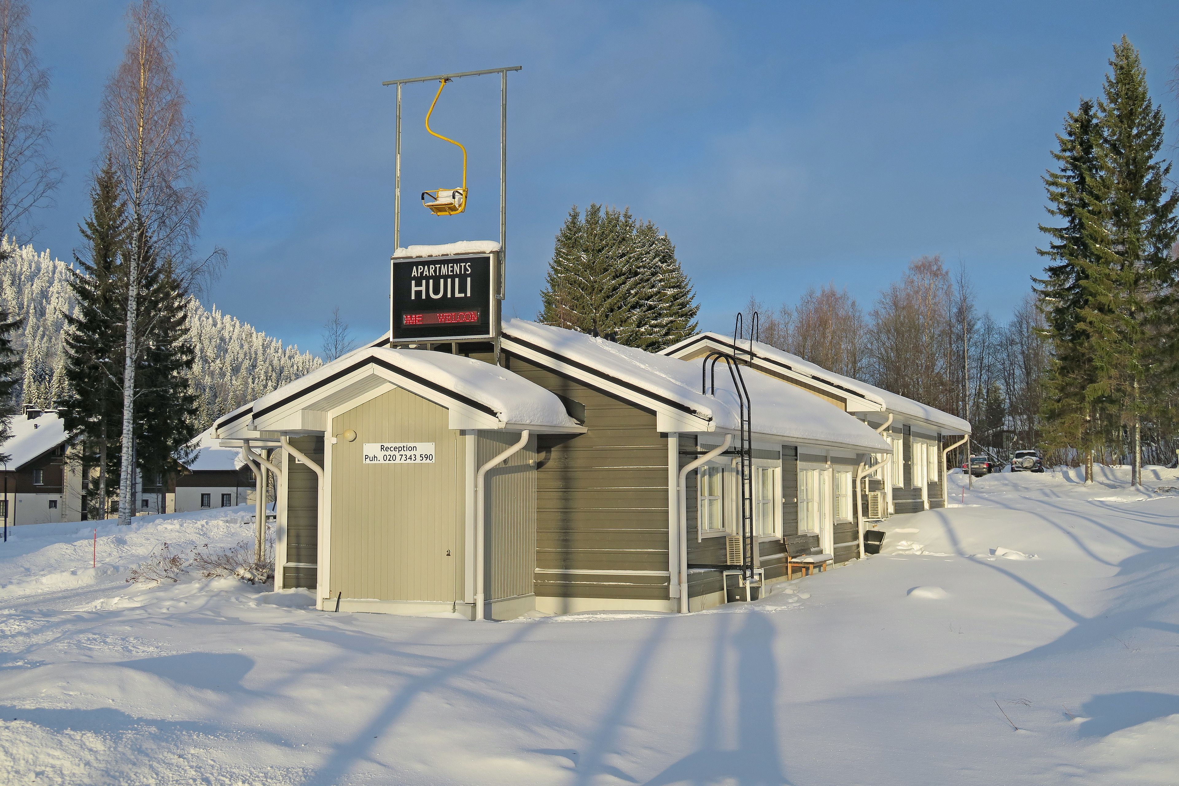 Un bâtiment enneigé, sur lequel on peut lire l’inscription « Appartements Huili », et une télécabine.