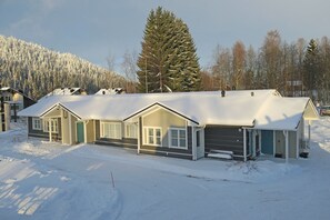 Une maison à deux étages, dont le toit est recouvert de neige, avec un grand arbre à feuilles persistantes en arrière-plan.
