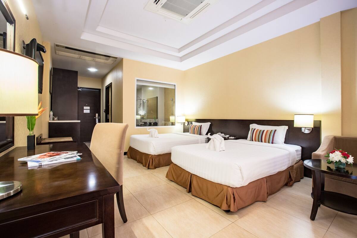 Quarto de hotel com duas camas, uma escrivaninha, uma cadeira, uma mesa com flores e um espelho.