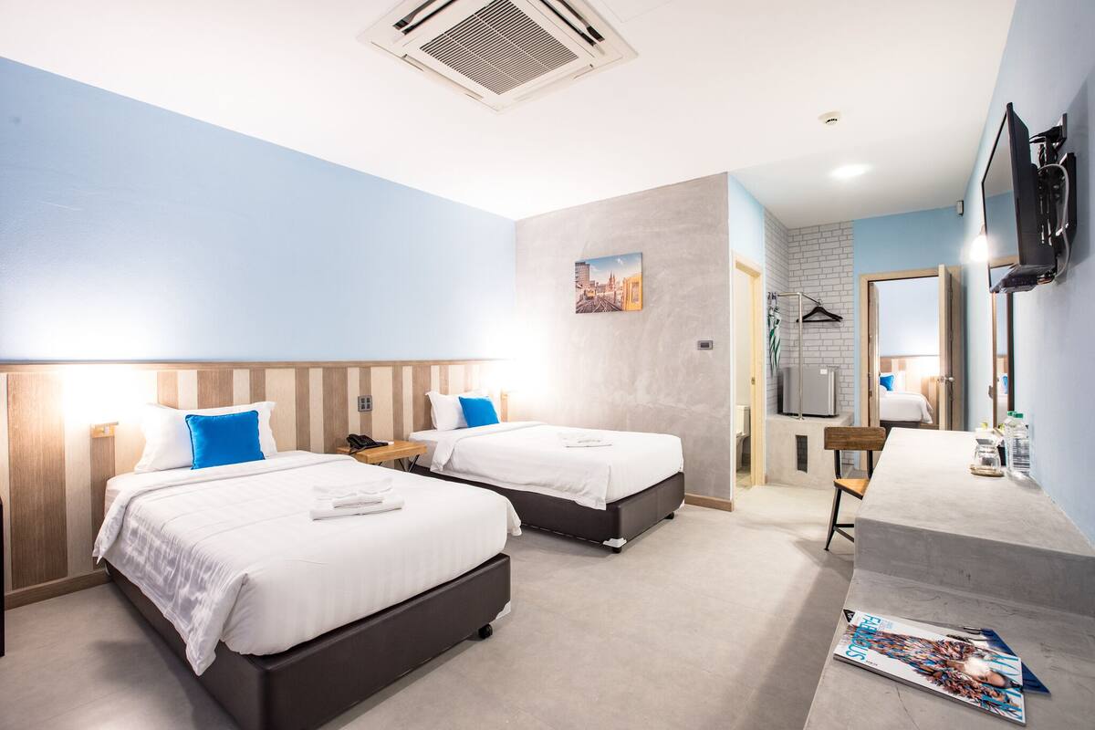 Quarto de hotel com duas camas, televisão, escrivaninha e banheiro visível através de uma porta aberta.