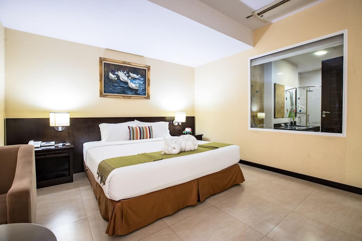 Quarto de hotel com uma cama grande, uma mesa de cabeceira com abajur, uma cadeira e um quadro na parede.
