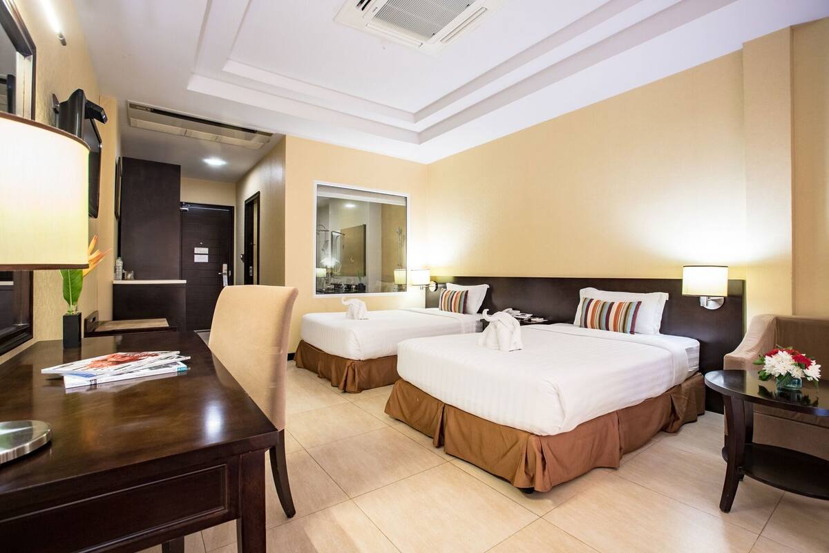 Quarto de hotel com duas camas, uma escrivaninha, uma cadeira, um espelho e uma mesinha de centro com um vaso de flores.