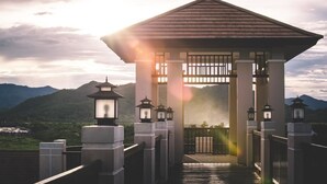 A traditional wooden pavilion with a tiled roof, surrounded by stone lanterns, set against a mountainous backdrop.