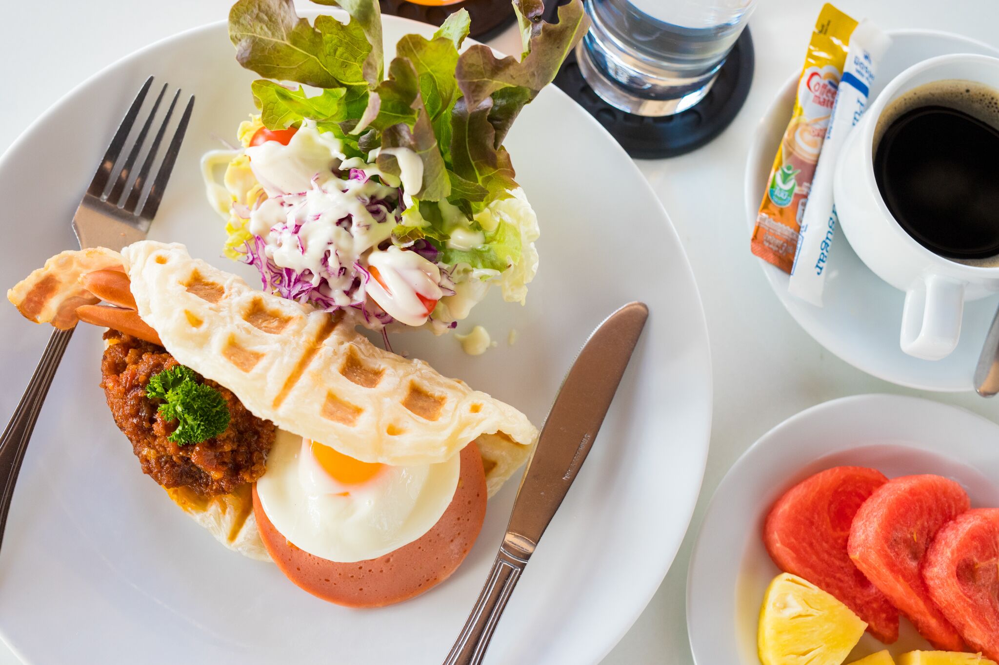 Un plato de desayuno con un gofre, huevo, albóndiga de carne y ensalada, una taza de café y un tazón de fruta.