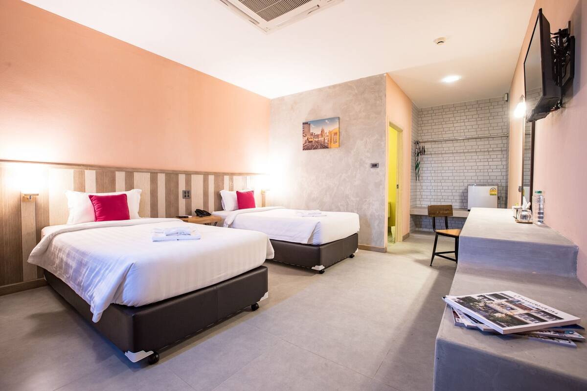 Quarto de hotel com duas camas, uma escrivaninha e um banheiro visível por uma porta aberta.