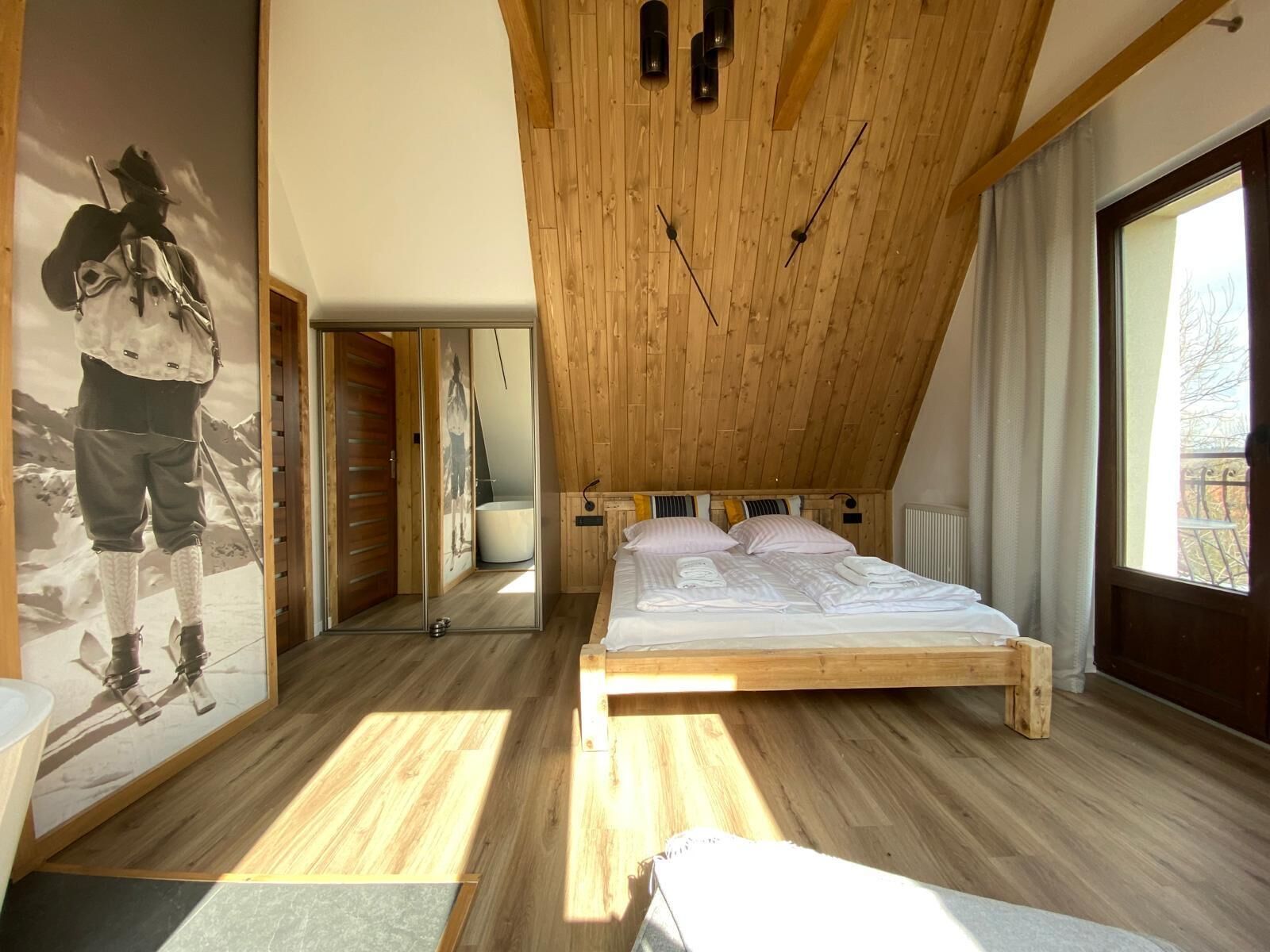 Een slaapkamer met een houten plafond, een bed met witte lakens, een groot raam en een spiegel aan de muur.