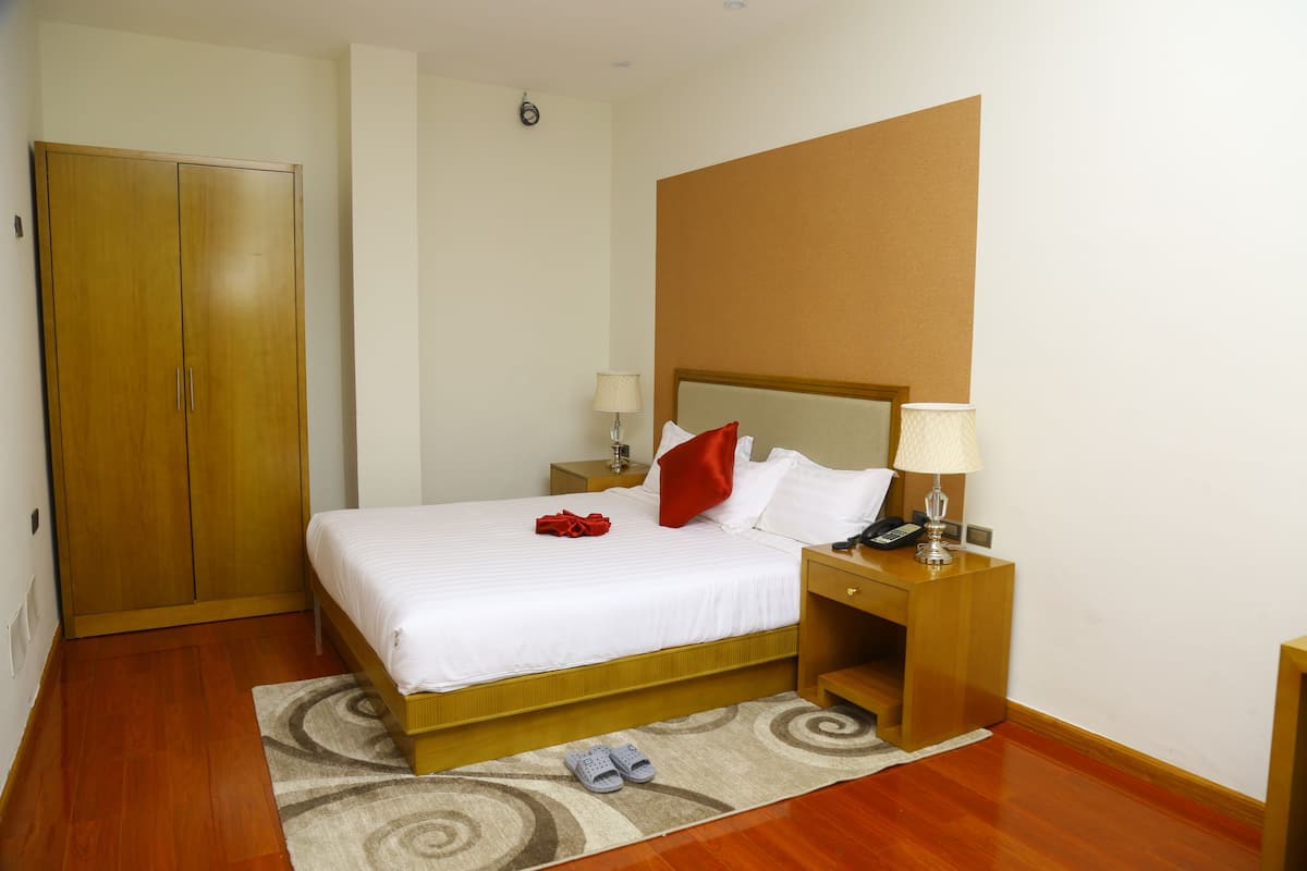 A hotel room with a wooden wardrobe, a bed with white bedding, a red pillow, a nightstand with a lamp, and a pair of slippers on the floor.