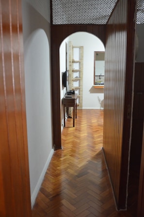 Un couloir doté d’un plancher en bois, d’un chambranle de porte en bois, et donnant sur une pièce où l’on aperçoit un bureau, des étagères et un miroir.