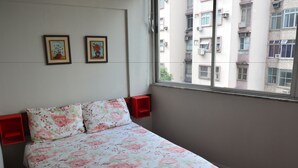 A bedroom with a floral bedspread, two wall-mounted pictures, and a window with a view of buildings. - Copacabana Etrusco Apartment (Rio de Janeiro)