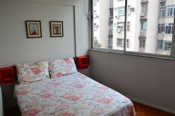 Une chambre à coucher avec une couvre-lit à motifs floraux, deux tableaux accrochés au mur et une fenêtre donnant sur des immeubles.