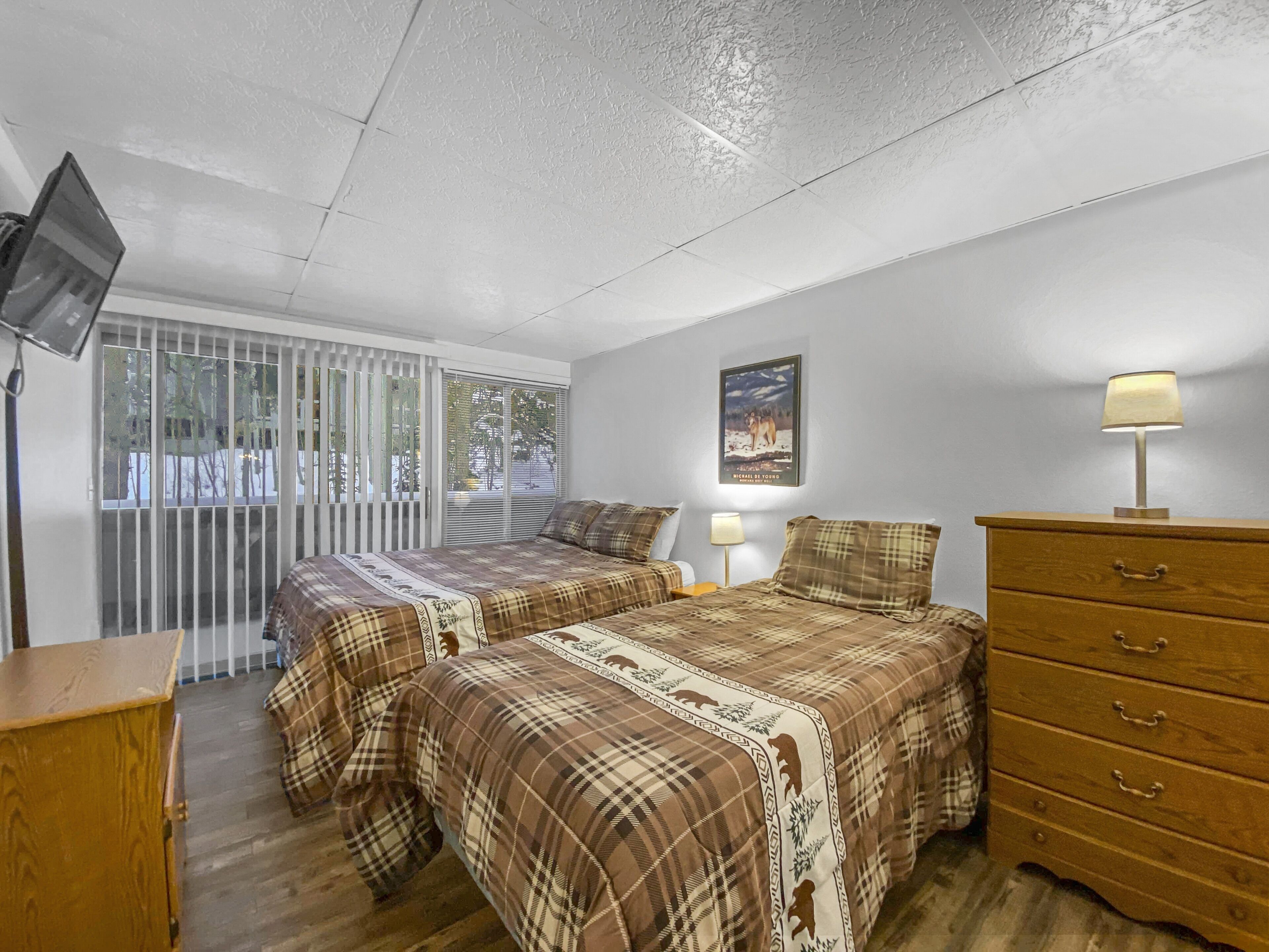 Quarto com duas camas, uma cômoda de madeira, televisão e uma janela com persianas.