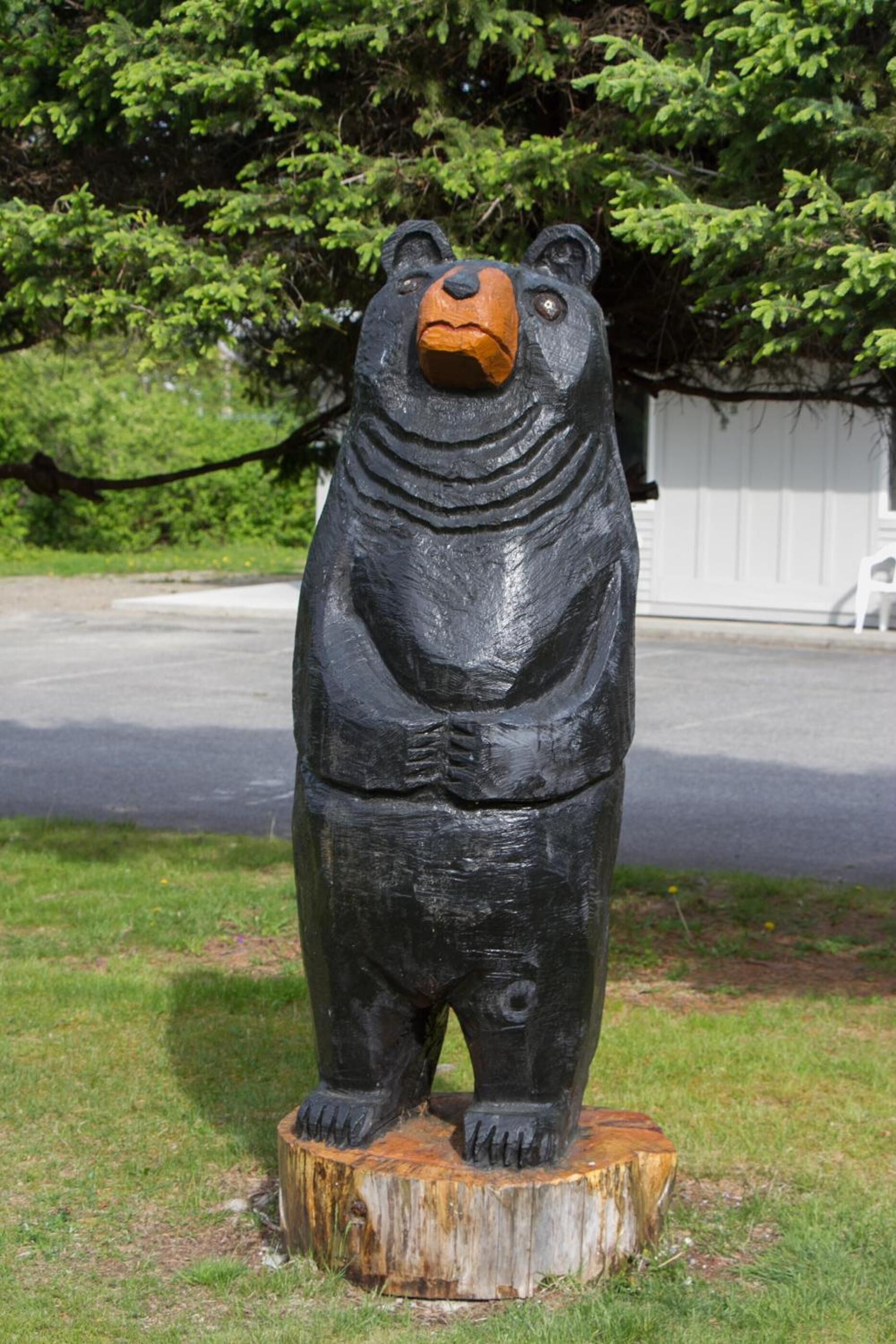 A black bear statue on a wooden stump in a grassy area with trees and a white fence in the background.