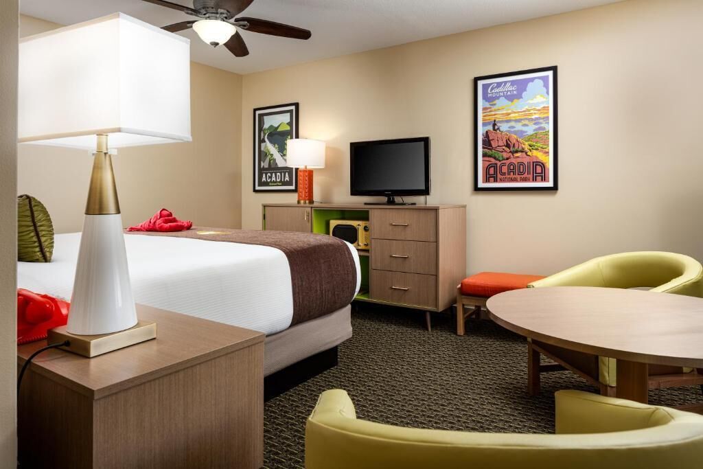 A hotel room with a bed, a desk with a lamp, a television, a chair, a table, and two posters on the wall.