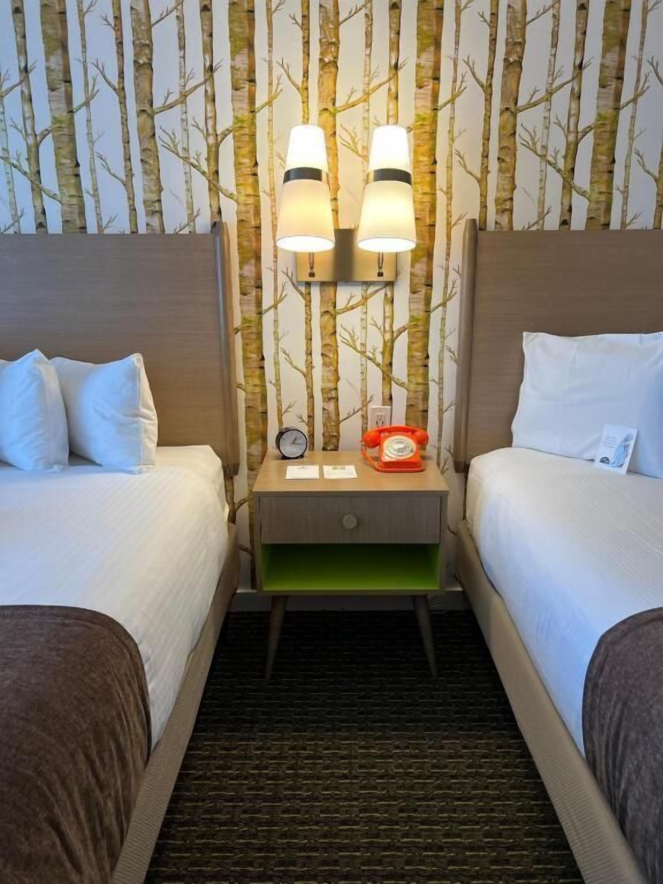 A hotel room with two beds, a nightstand with a clock and a red telephone, and wall-mounted lamps.